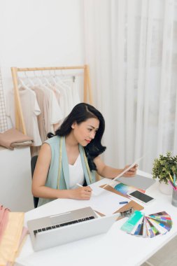 Genç Asyalı moda tasarımcısının portresi atölyede dizüstü bilgisayarının önünde oturuyor.