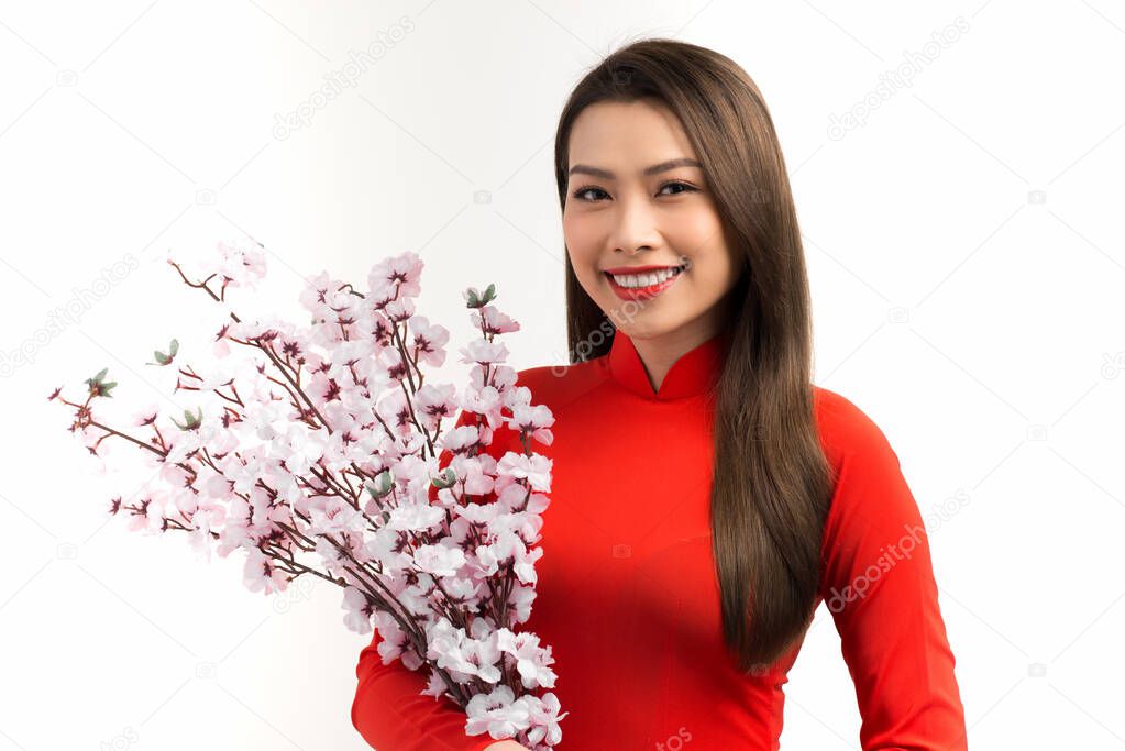 Elegant Asian woman holding peach flowers while wearing Ao dai over white background.