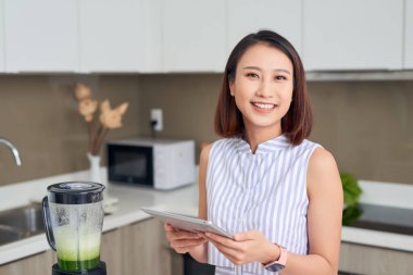 Asyalı bir kadın mutfakta meyve ve sebzeli smoothie yapmak için tablet kullanıyor..