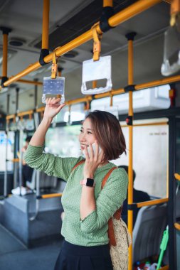 Genç sevimli neşeli kadın telefonu kullanarak otobüste duruyor ve gülümsüyor.
