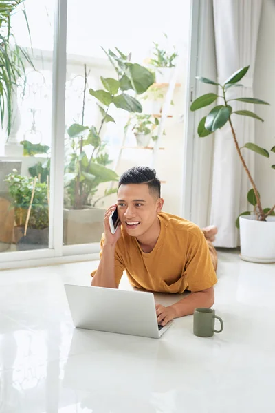 Sorrindo Homem Falando Celular Usando Laptop Enquanto Deitado Chão Casa — Fotografia de Stock