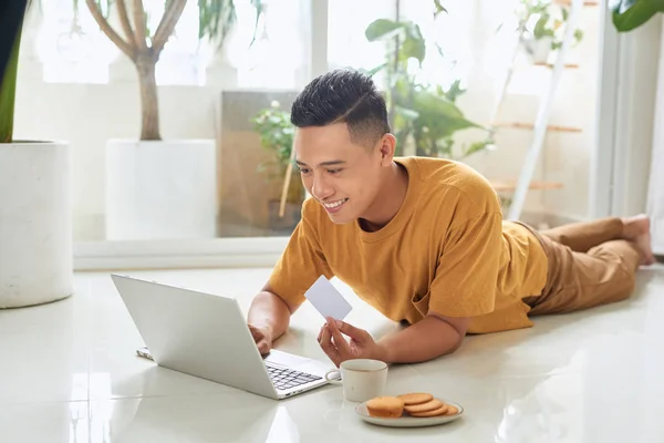 英俊的年轻人拿着信用卡 微笑着在地板上使用笔记本电脑 — 图库照片