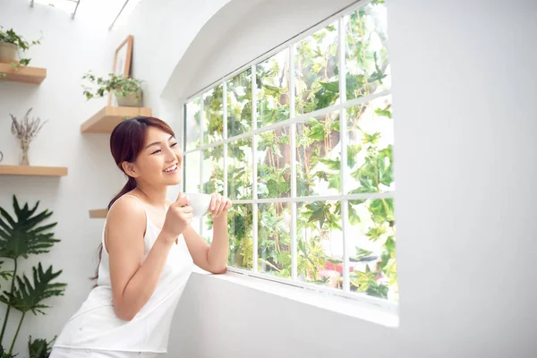 Pencerenin arkasında duran, kahve içen ve dışarı bakan güzel bir kadın dinlenmenin keyfini çıkarıyor.