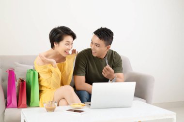 Evdeki masada otururken internetten bilgisayarla alışveriş yapan neşeli bir çiftin portresi.