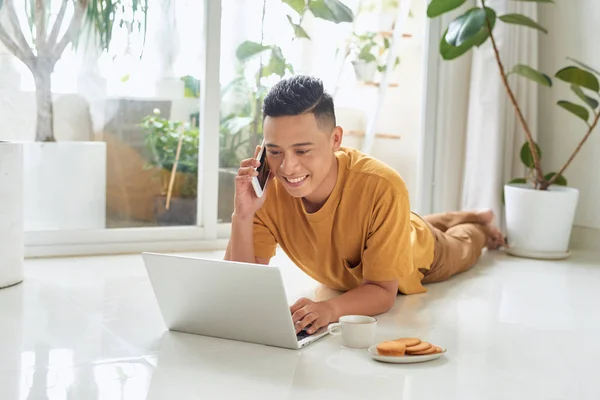 Sorrindo Homem Falando Celular Usando Laptop Enquanto Deitado Chão Casa — Fotografia de Stock