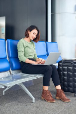Havaalanı terminalinde dizüstü bilgisayar kullanan Asyalı kadın seyahat için valiziyle oturuyor.