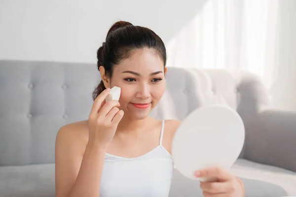 Beautiful asian woman using cosmetic sponge on face and look mirror at home