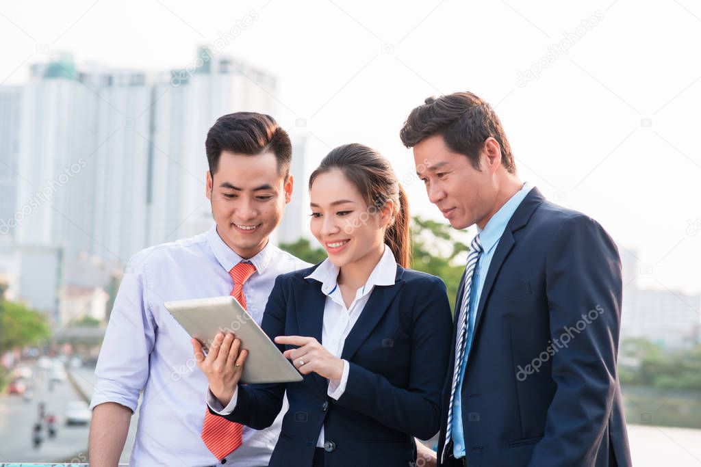Asian businessman in suit showing online presentation on digital tablet to his colleagues while they standing