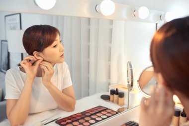 Genç Asyalı kadın aynanın önünde küpe takıyor.