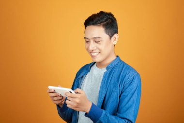 Parlak turuncu stüdyo arka planında izole edilmiş akıllı telefon kullanan gülümseyen yakışıklı Asyalı adam.