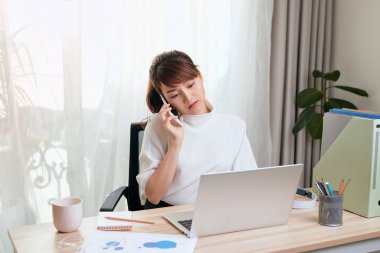 Asyalı Kadın telefonda konuşurken ofiste dizüstü bilgisayarla çalışıyor.