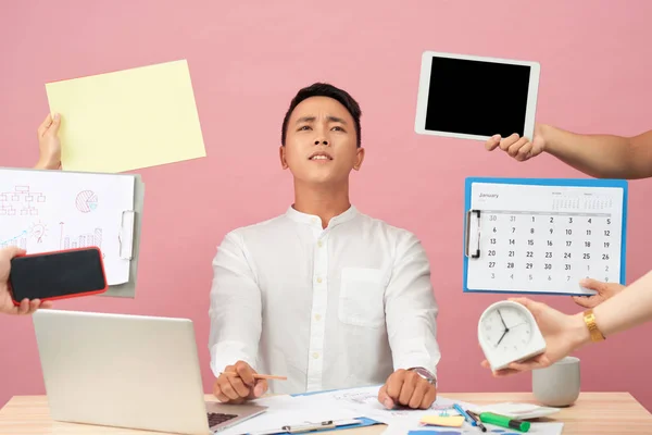 Sad young man sits at desktop, hands with papers, alarm clock, touchpad, notepad with stickers, studies documentation, isolated over pink background