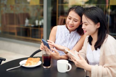 Kahve molası veren iki genç kadın akıllı telefon kullanıyor. Kaldırımdaki kafede cep telefonu kullanan mutlu kadınlar.