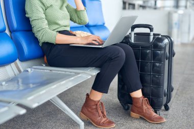 Havaalanı terminalinde dizüstü bilgisayar kullanan Asyalı kadın seyahat için valiziyle oturuyor.