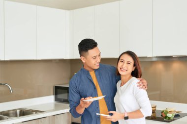 Heyecanlı neşeli genç çift mutfakta otururken sağlıklı salata pişirme, cep telefonu bakarak