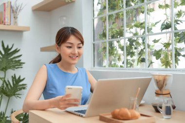 Pencerenin arkasında çalışırken telefon ve laptop kullanan mutlu genç Asyalı kadın..