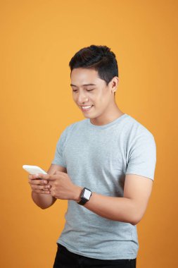 Akıllı telefon kullanan yakışıklı Asyalı genç adam, stüdyonun turuncu arka planında vuruldu.