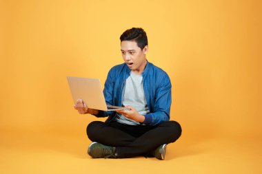 Yerde oturan genç adam turuncu arka planda izole edilmiş dizüstü bilgisayarla çalışıyor..