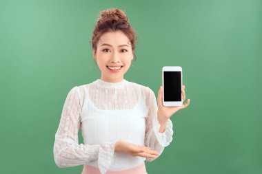 Boş cep telefonunu gösteren Asyalı genç bir kadının portresi. Yeşil arkaplan üzerinde izole edilmiş.