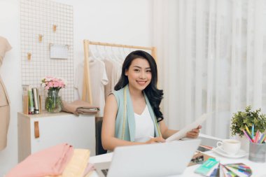 Günlük giysiler içinde genç Asyalı mutlu bir tasarımcı masa başında oturmuş moda skeçleri yapıyor.