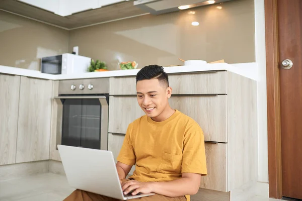 细心的亚洲男子使用笔记本电脑 而坐在厨房的地板上 — 图库照片