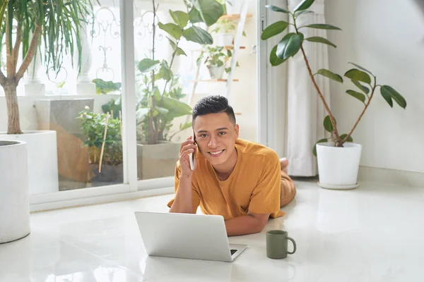 Sorrindo Homem Falando Celular Usando Laptop Enquanto Deitado Chão Casa — Fotografia de Stock