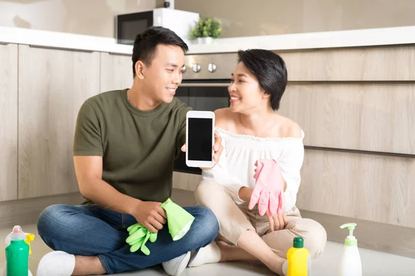 Casal Jovem Fazendo Tarefas Domésticas Enquanto Mostra Telefone Celular Cozinha — Fotografia de Stock