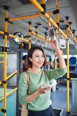 Genç sevimli neşeli kadın telefonu kullanarak otobüste duruyor ve gülümsüyor.