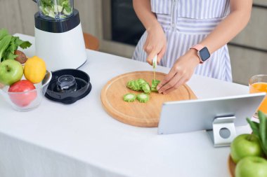 Asyalı kadın önünde tabletle mutfakta sebze kesiyor.