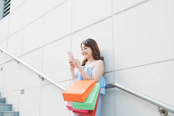 兴高采烈的亚洲女人一边站在墙后一边拿着五颜六色的袋子 一边用手机 购物概念 — 图库照片