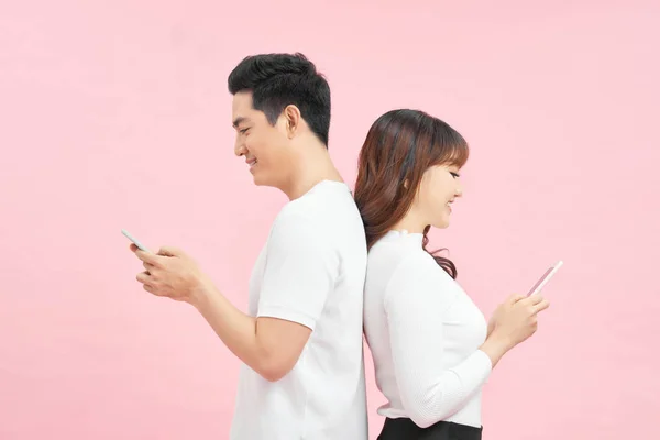 若いカップルはピンクの背景に立っている間にスマートフォンや笑顔を使用しています — ストック写真