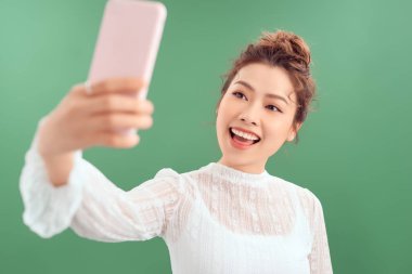 Havalı neşeli kızın portresi elinde akıllı bir telefonla yeşil arka planda izole edilmiş selfie çekiyor.