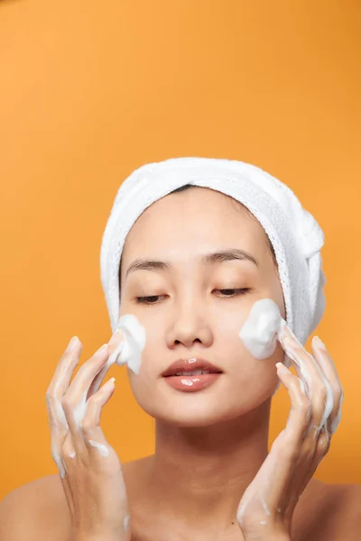 年轻的亚洲女人穿着浴巾 在脸上涂奶油 在脸上触摸 在橙色背景下分离 — 图库照片