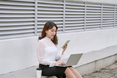 güneşli bir gün boyunca sokak yapraklı bir şehirde otururken bir dizüstü bilgisayar kullanarak cazip iş kadını gülümseyen.