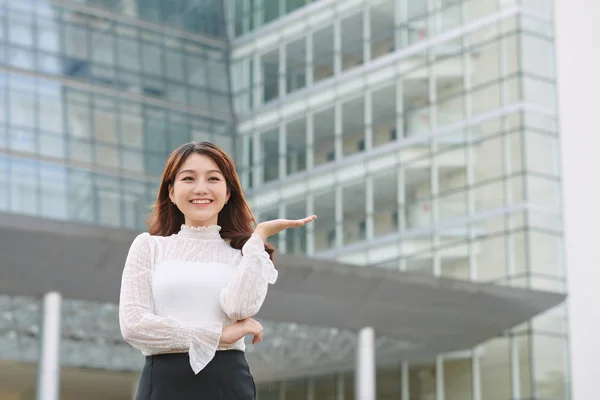 Beautiful asian woman with friendly smile has optimistic expression, shows something with her hand opened, standing outdoors. Youth and advertisement concept.