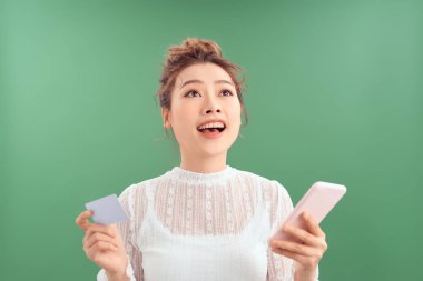 Yeşil arka planda kredi kartı ve cep telefonu tutan gülümseyen Asyalı kızın portresi.