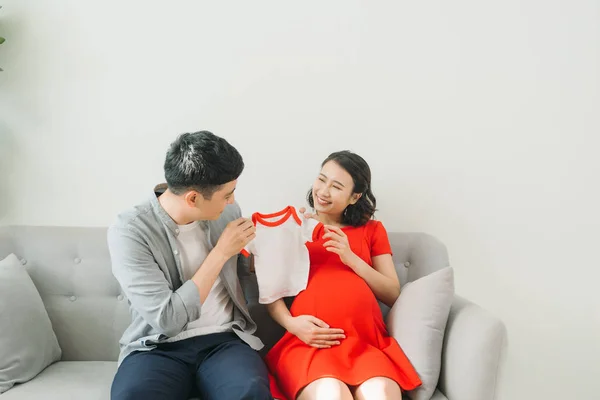 幸せな妊婦保持赤ちゃんTシャツと彼女の夫の家でソファの上に — ストック写真