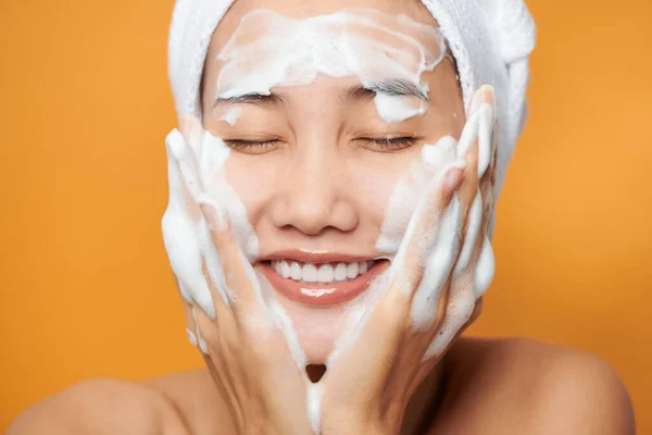 バスタオルで顔にクリームを塗り 顔に触れるアジア系の若い女性 オレンジの背景に隔離された — ストック写真