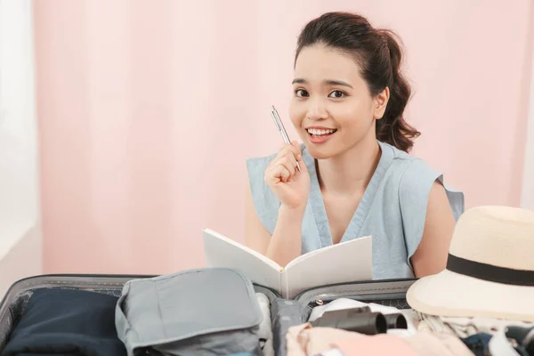 It is better to write everything! Young woman in a bright room in front of clothes thinks what else needs to be folded into a suitcase before traveling