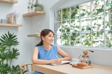 Genç Asyalı kadın evde çalışıyor, pencerenin arkasındaki masaya kağıt okuyor ve yazıyor.