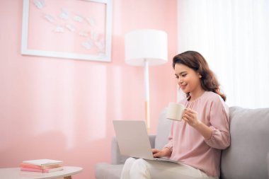 Çekici Asyalı kadın yaşam tarzı dizüstü bilgisayarını kullanarak kanepede oturup kahve içiyor.