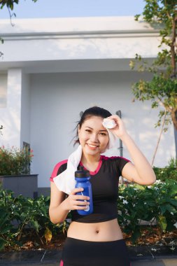Göz kamaştırıcı fitness Asyalı kadın. Koşudan sonra terliyor ve bir şişe su tutuyor. Ya da dışarıda koşuyor.