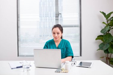 Hastane ofisinde dizüstü bilgisayarla yazı yazan genç bir kadın doktor..