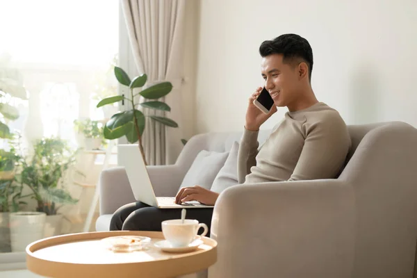 Jovem Sentado Sofá Casa Com Laptop Conversando Com Amigo Smartphone — Fotografia de Stock