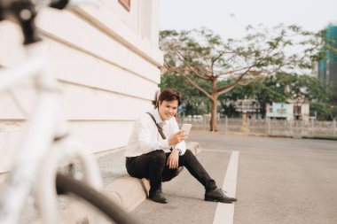 Dijital tablet kullanan, neşeli bir şekilde gülümseyen, komik bir şey okuyan, yerde, bisikletin yanında, dışarıda oturan modaya uyan genç sakallı bir adam..