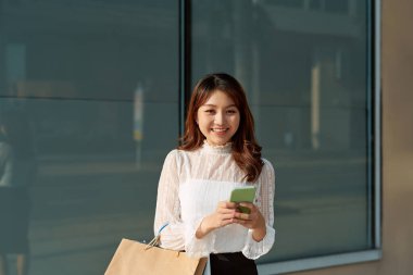 Vitrin kenarında alışveriş torbaları olan güzel bir moda kadını akıllı telefonları arıyor.