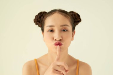 Keep in secret! Portrait of playful positive Asian teen girl with buns hairstyle showing silence gesture over light background. clipart