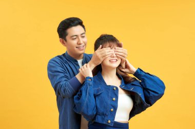 Guess who. Playful young guy standing behind girlfriend and closing her eyes, yellow studio background with copy space clipart