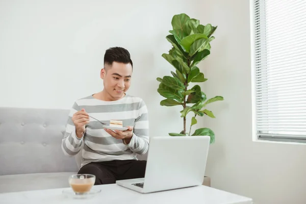 Jovem Asiático Comendo Bolo Quando Sentado Sofá Usando Laptop Trabalho — Fotografia de Stock