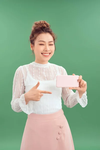 幸せな若いアジアの女性は彼女のスマートフォンを指しながら 緑の背景に隔離された立って — ストック写真